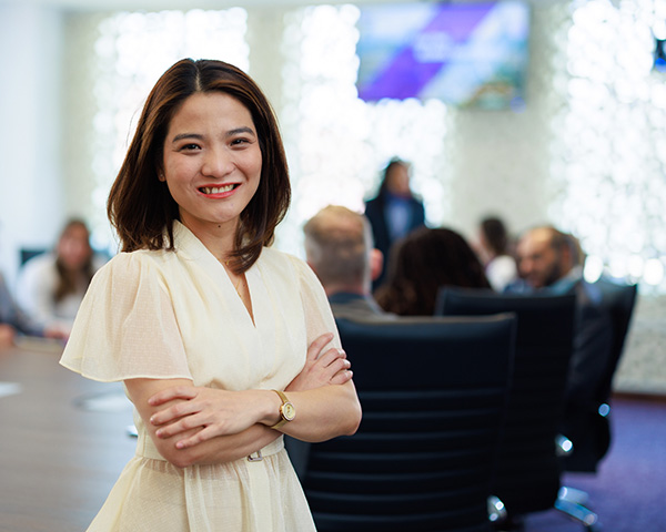 MBA student stands, smiling, as others gather around the boardroom table