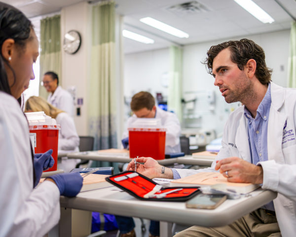 Two medical students in white coats, one sitting at a desk, and the other looking at surgical instruments