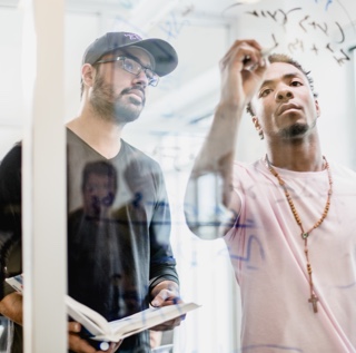 TCU Students Writing on a Glass Partition One male student holding a book looks at what another male student is writing on a glass wall