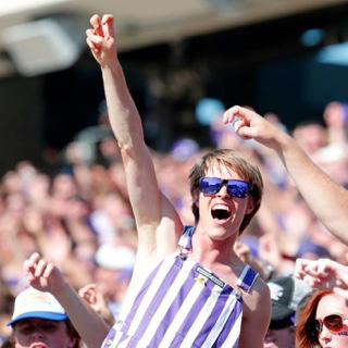 A happy, cheering student in striped overall and purple sunglasses makes the two-fingered "Go Frogs" hand sign at a crowded football game.