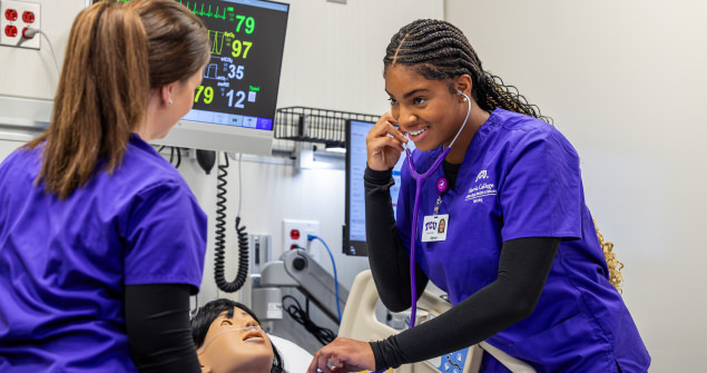 A nursing student in purple scrubs uses a stethoscope on a simulation manekin