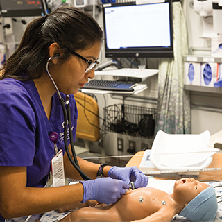 A nursing student in purple scrubs uses a stethoscope as she practices on an infant simulation dummy