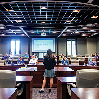 A young woman in a suit speaks before a room full of TCU Honors professors