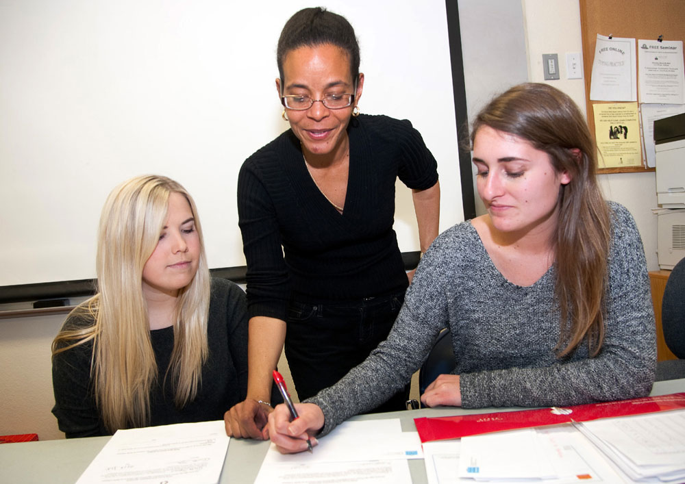 An economics professors looks on as one of two students writes out an idea on a piece of paper