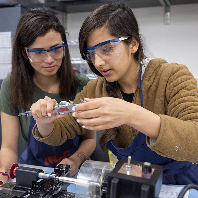 Engineering students carefully measure a machined part using a dial caliper