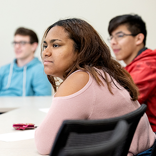 Three TCU students of different ethnicities listen together in a classroom. A young woman in a pink sweater is in the foreground.