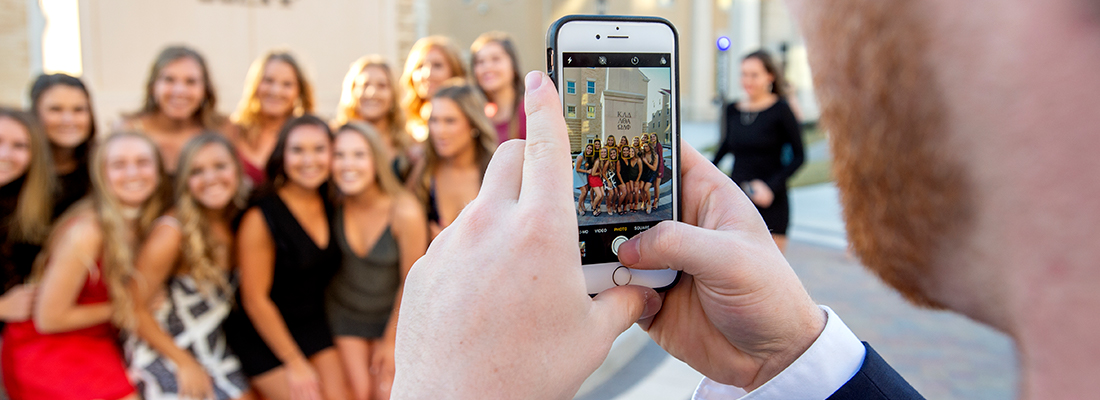 A young man uses a cell phone to photograph a group of smiling sorority girls before a party in the Greek Village at TCU.